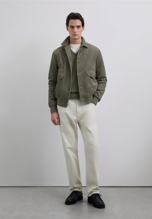 Olijfgroene corduroy jas met twee voorzakken, gedragen over een groene trui, gecombineerd met witte broek en zwarte schoenen. Neutrale achtergrond.