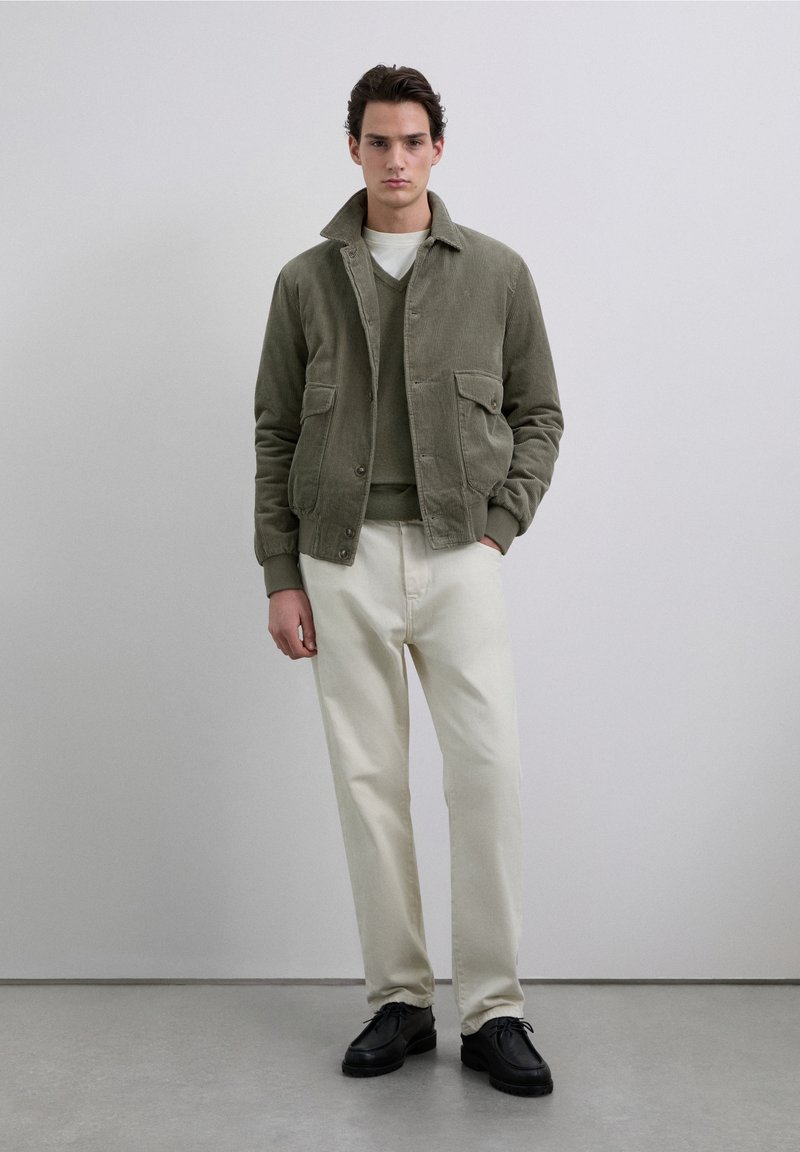 Chaqueta de pana color oliva con dos bolsillos delanteros, puesta sobre un suéter verde, combinada con pantalones blancos y zapatos negros. Fondo neutro.