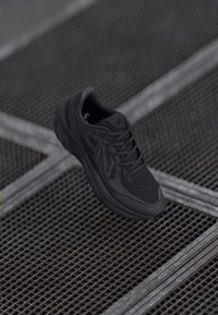 Chaussure de sport noire avec un matériau en maille respirante, un col rembourré, des détails texturés et un marquage visible sur le côté, reposant sur une surface en grille.