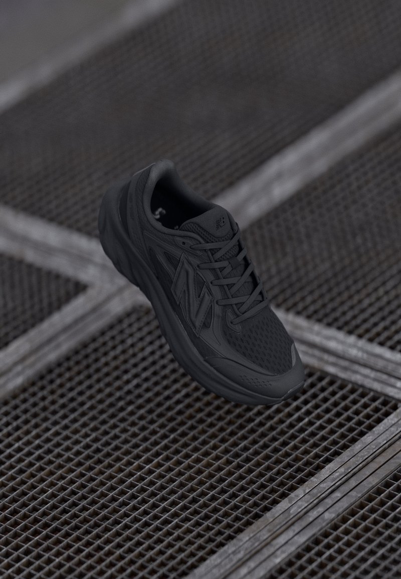 Chaussure de sport noire avec un matériau en maille respirante, un col rembourré, des détails texturés et un marquage visible sur le côté, reposant sur une surface en grille.