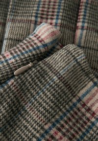 Wollen stof met een plaidpatroon in grijs, blauw en rode tinten, met een close-up weergave die de textuur en een klein ritsdetail laat zien.
