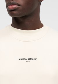 Gros plan sur une personne portant un sweat-shirt couleur crème avec l'inscription "MAISON KITSUNÉ PARIS" en noir sur la poitrine.