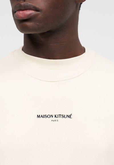 Κοντινό πλάνο ενός ατόμου που φοράει φούτερ σε κρεμ απόχρωση με την επιγραφή «MAISON KITSUNÉ PARIS» τυπωμένη με μαύρα γράμματα στο στήθος.