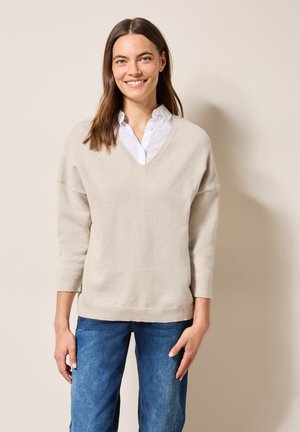 Hellbeige V-Ausschnitt-Pullover mit lockerer Passform, 3/4-Ärmeln und gerippten Bündchen. Über einem weißen Hemd mit Kragen getragen. Denim-Jeans blitzen darunter hervor.
