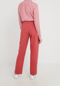 Osoba stojící zády k divákovi, oblečená v červených širokých kalhotách, dlouhém tričku s červenými a bílými pruhy a bílých teniskách.