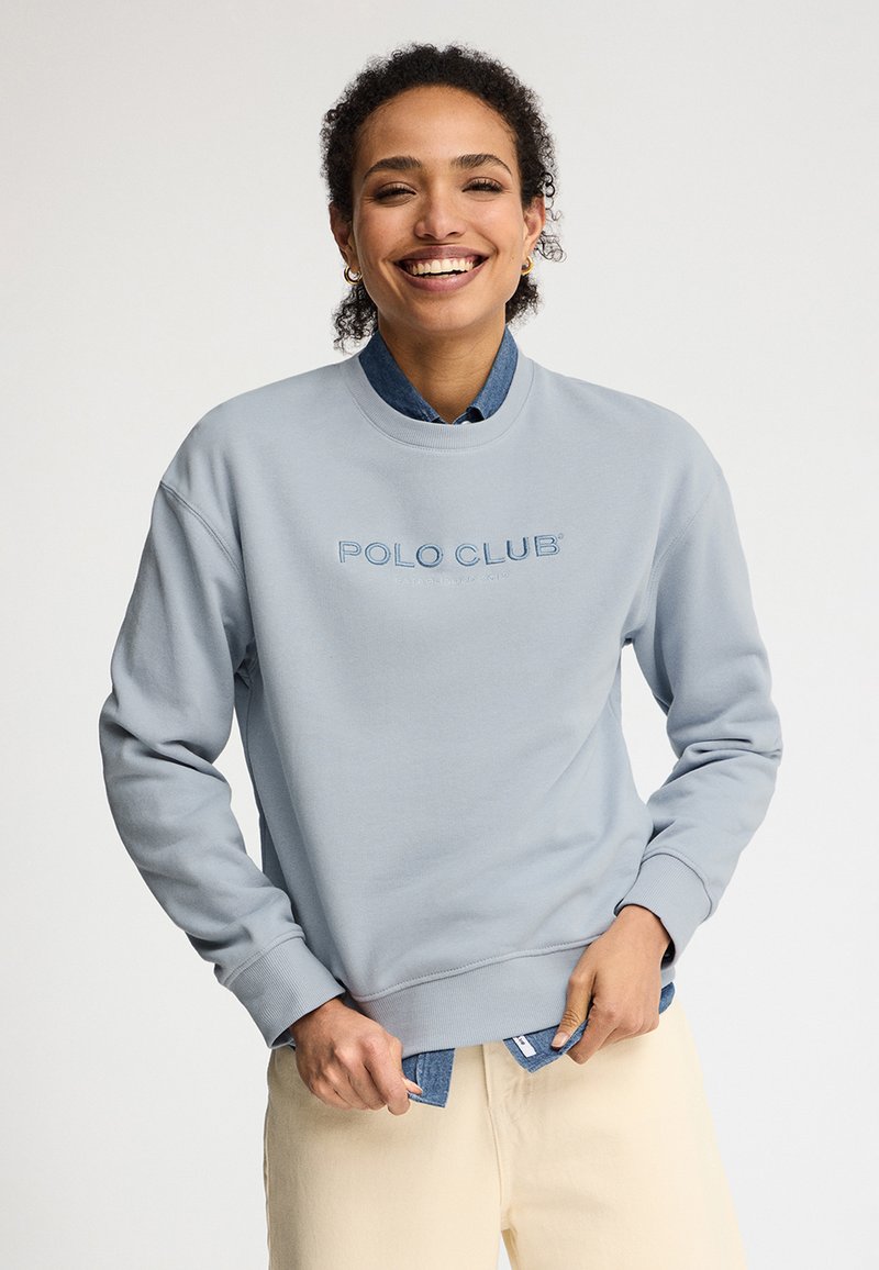 Persona sorridente che indossa una felpa azzurro chiaro con la scritta "POLO CLUB" sopra una camicia di jeans e pantaloni beige, che sistema l'orlo della camicia su uno sfondo semplice.
