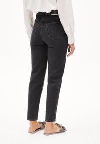 Person som bär svarta högmidjade jeans med bakfickor och en vit skjorta, står barfota i gråa slip-in sandaler med rosett.