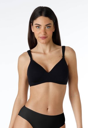 Donna con capelli lunghi e scuri che indossa un reggiseno da allattamento nero e biancheria intima coordinata, in piedi davanti a uno sfondo bianco uniforme.