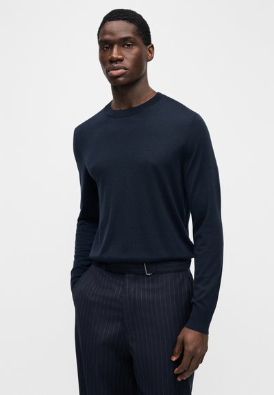 Πλεκτό πουλόβερ μακρυμάνικο με στρογγυλή λαιμόκοψη σε ναυτικό μπλε με διακριτικό λογότυπο. Φορεμένο με παντελόνια με λεπτές ρίγες, που αναδεικνύουν υφή λεία και εφαρμοστή γραμμή.