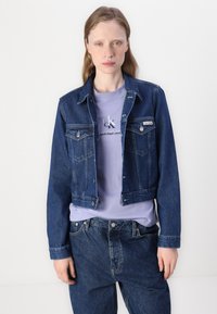 Giacca di jeans blu scuro con design cropped, caratterizzata da chiusure a bottoni, tasche sul petto e cuciture a contrasto sopra una camicia lilla.
