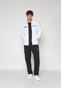 Hvit hettegenser med glidelås og svart "Champion" logo, lagd over en svart t-skjorte, sammen med svarte joggebukser og hvite joggesko.