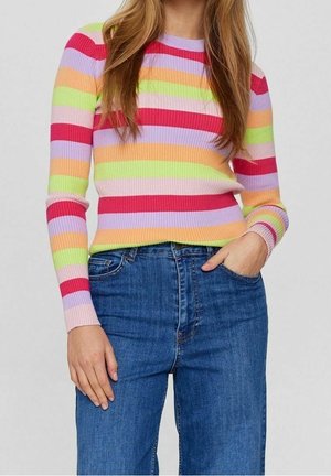 Personne portant un pull côtelé à rayures horizontales rouges, roses, violettes, orange et vertes, associé à un jean bleu, main dans la poche.