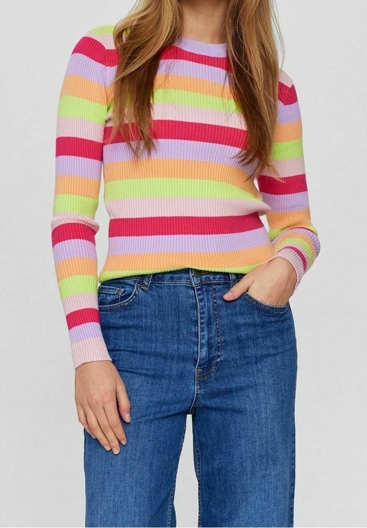 Personne portant un pull côtelé à rayures horizontales rouges, roses, violettes, orange et vertes, associé à un jean bleu, main dans la poche.
