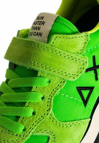 Sneaker verde con una combinazione di materiali in suede e tessuto, una cinturino in Velcro, dettagli neri e un'etichetta testuale.
