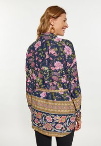 Blusa floreale a maniche lunghe in blu navy con fiori rosa e verdi. Presenta un orlo accentato in oro e una vita cintrata per una vestibilità strutturata.