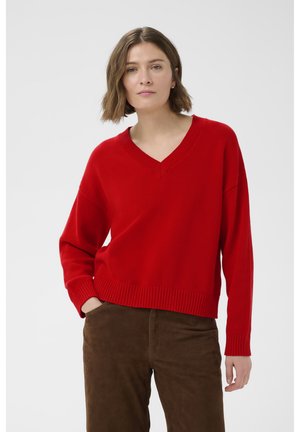Kvinde med kort brunt hår iført en rød V-hals trøje og brune bukser, stående med den ene hånd i lommen mod en hvid baggrund.