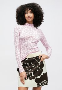 Ženska z kodrastimi lasmi, oblečena v rožnat dolg rokav z vzorcem cvetja in kremno krilo z velikimi črnimi cvetličnimi motivi, pozira pred enobarvnim ozadjem.