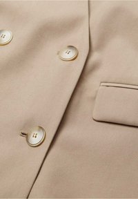Beige Jackenstoff mit glatter Textur, ausgestattet mit zwei runden, durchsichtigen Knöpfen und einer kleinen Brusttasche auf der linken Seite.
