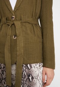 Olivově zelený lněný blazer s páskem v pase, dvěma předními kapsami a hnědými knoflíky, spárovaný s kalhotami s hadím vzorem.