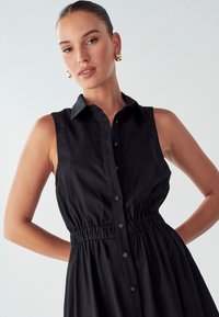 Juodas be rankovių suknelė su mygtukų užsegimu priekyje, apykakle ir surinkta talija. Audinys atrodo lygus su subtiliu blizgesiu.