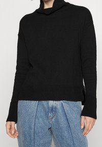 Black knit sweater with a high cowl neck, ribbed texture, and side slits, paired with light blue loose-fitting jeans.
