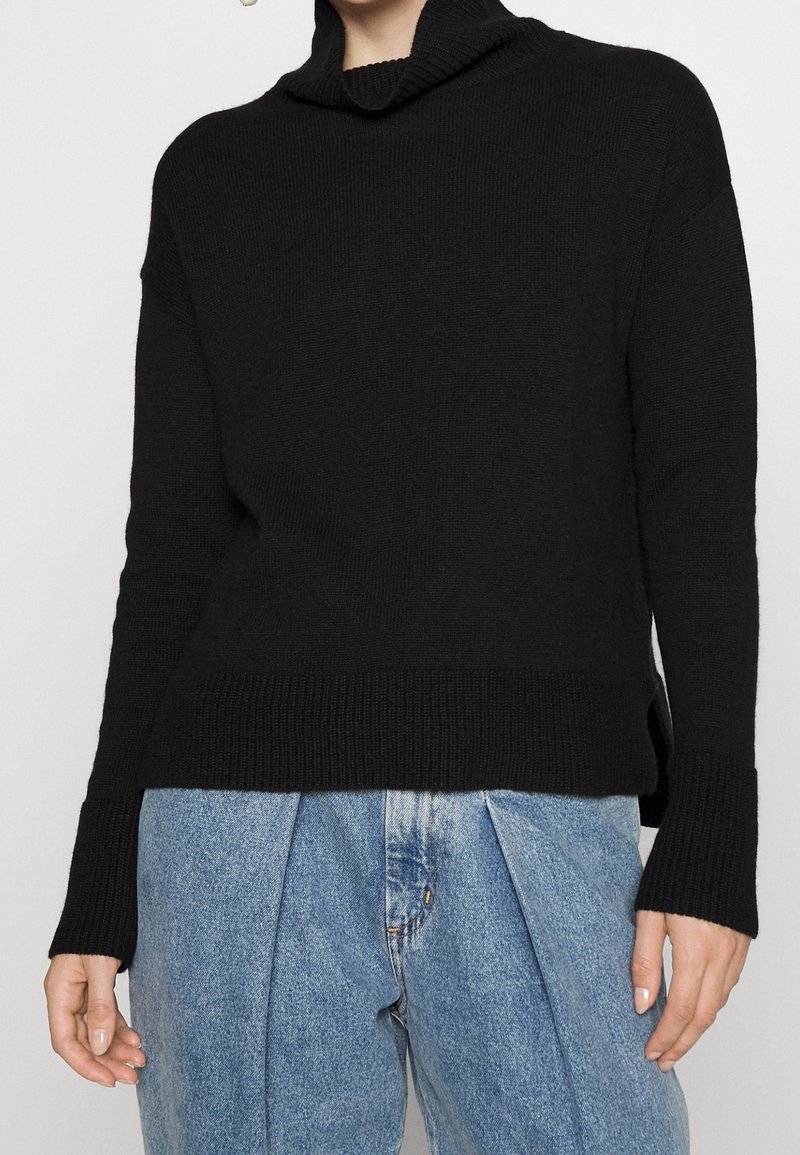 Black knit sweater with a high cowl neck, ribbed texture, and side slits, paired with light blue loose-fitting jeans.