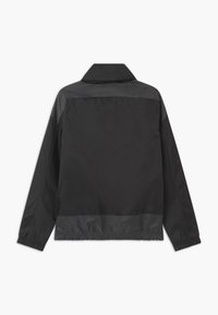 Mustat kevyet takki, jossa on kauluksellinen kaula-aukko ja joustava vyötärö. Sujuva pinta, jossa on kontrastiväriset matta- ja kiiltopaneelit harteilla.