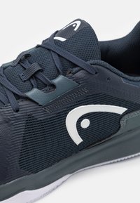 Σκούρο μπλε αθλητικό παπούτσι από πλέγμα και καουτσούκ, που διαθέτει λευκό λογότυπο, υφασμάτινες λεπτομέρειες και χαμηλό προφίλ.