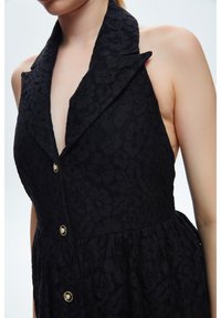 Abito in pizzo nero con scollatura a V profonda, colletto strutturato e cinque bottoni decorativi. Il tessuto presenta un motivo floreale con una finitura texture.