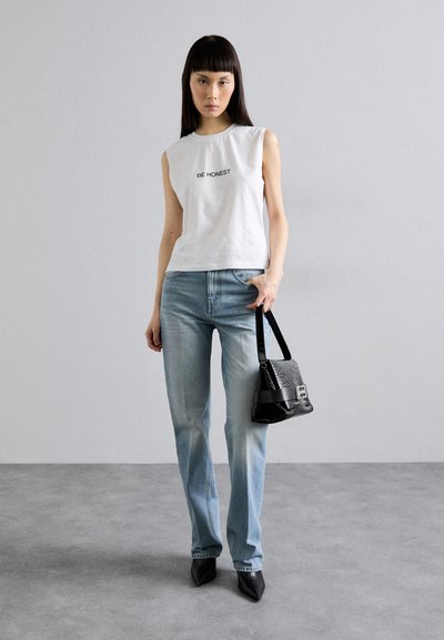 Weißes ärmelloses Oberteil mit dem Text "SEI EHRLICH", hellblaue Jeans, schwarze Handtasche mit strukturierter Oberfläche und spitze schwarze Absätze.