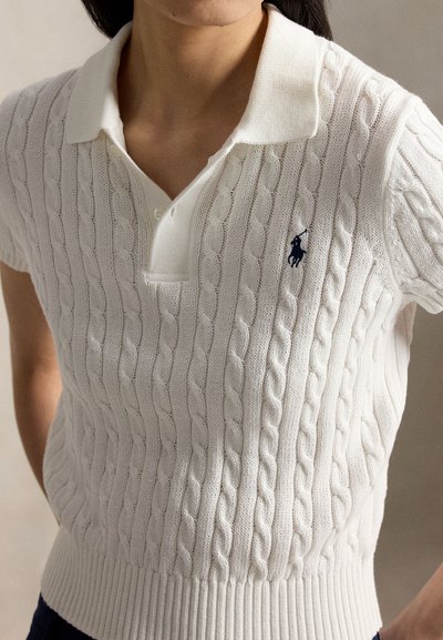 Person wearing a white short-sleeve cable-knit polo shirt with a dark embroidered logo on the left chest.