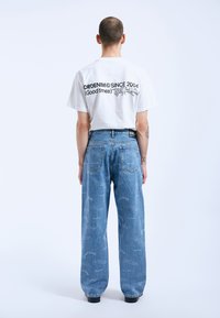 Camiseta blanca con texto gráfico negro en la parte posterior, combinada con jeans azules holgados que presentan un patrón repetido en azul claro.