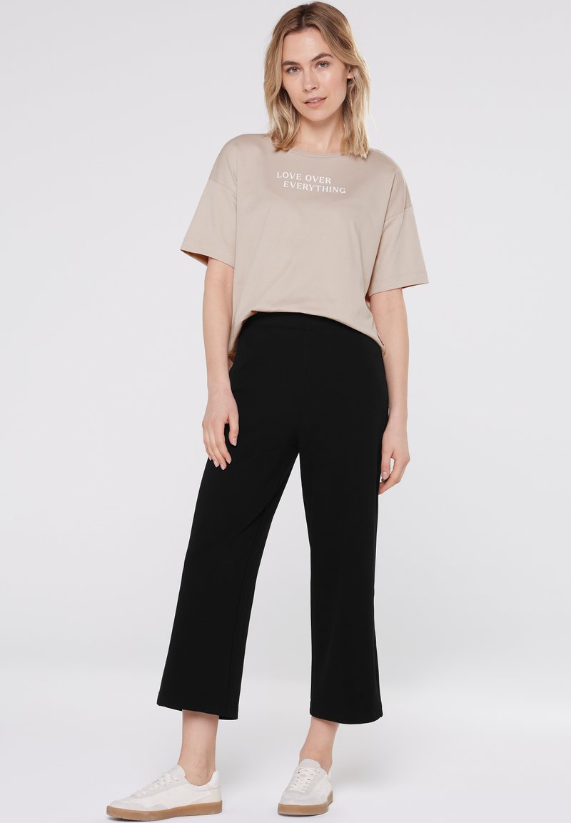 Frau in beiger T-Shirt mit dem Aufdruck "LOVE OVER EVERYTHING", schwarzen weiten Hosen und weißen Sneakern, stehend vor einem einfarbigen Hintergrund.