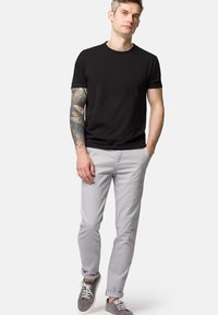 Czarny t-shirt z krótkim rękawem, szare spodnie slim-fit, szare buty. Zauważalny tatuaż na prawym ramieniu. Neutralne tło.
