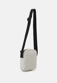 Piccola borsa a tracolla di colore grigio chiaro con una tracolla in tessuto nero regolabile e chiusura superiore con zip, mostrata su uno sfondo bianco.