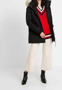 Svart parkas med pälsfodrad huva, ovanpå en röd V-ringad tröja med vita och marinblå ränder, kombinerad med beige vida byxor och svarta stövlar.