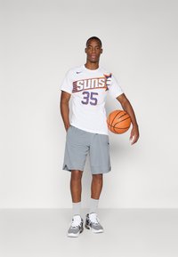 Maglietta sportiva bianca con logo "Suns" e numero 35, abbinata a pantaloni corti grigi chiari. Scarpe nere e bianche, mentre tiene un pallone da basket.