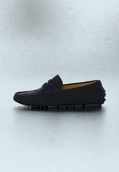 Μαύρο δερμάτινο μοκασίνι με λαστιχένια σόλα με καρφιά και λεπτομέρεια λουριού τύπου penny, εμφανίζεται από το πλάι σε ανοιχτό γκρι επιφάνεια.