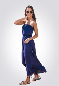 Jumpsuit blu navy senza spalline con corpino smock, vita elasticizzata e orlo arricciato. Abbinato a sandali con cinturino e occhiali da sole.