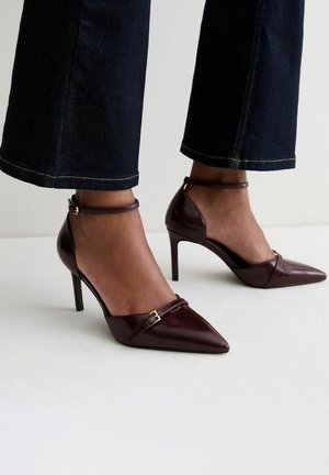 Tacones altos de punta fina de color marrón oscuro con tiras en el tobillo, usados con pantalones cortos azul oscuro sobre fondo claro.