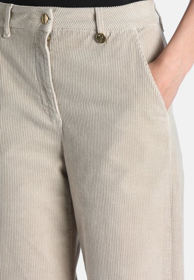 Gros plan sur un pantalon en velours côtelé beige avec une main dans la poche avant et des boutons métalliques décoratifs à la taille.