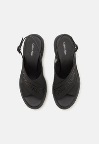 Černé sandály s gumovou podrážkou, které mají křížové elastické popruhy a na jednom popruhu je vyražený název značky "Calvin Klein".