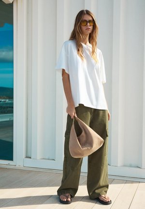 Femme portant un t-shirt blanc surdimensionné, un pantalon cargo ample olive, des lunettes de soleil teintées jaunes, tenant un sac à épaule beige, debout sur une terrasse en bois.