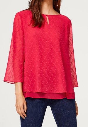 Kvinde iført en lys rosa tekstureret bluse med lange ærmer og en lille nøglehulshalsudskæring, kombineret med mørkeblå jeans.