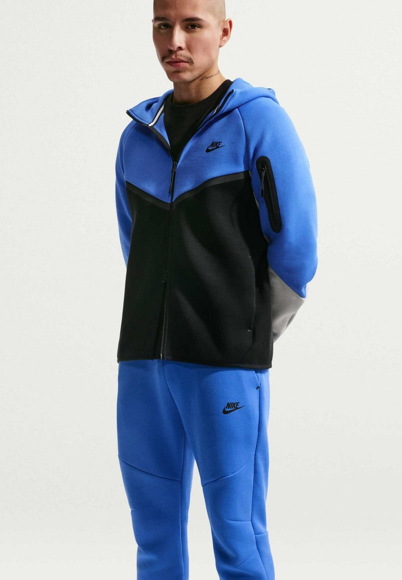 Homme portant un sweat à capuche zippé Nike bleu et noir et un pantalon de survêtement Nike bleu assorti sur fond blanc.