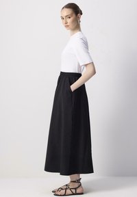 Jupe longue noire avec poches latérales, taille élastique, associée à un t-shirt blanc à col rond, portée avec des sandales noires à lanières.