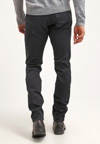 Vaqueros de mezclilla negra de corte ajustado con textura suave, con costuras en contraste en los bolsillos traseros y combinados con botas de cuero oscuro.