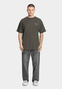 Ciemnozielona, luźna bawełniana koszulka z okrągłym dekoltem, z małym złotym logo. Połączona z jasnoszarymi jeansami i białymi butami.