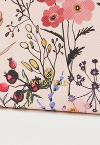 Portefeuille en tissu à motif floral dans un rose doux, présentant des fleurs et des feuilles vibrantes. Logo doré "ANNA FIELD" en accent près du bas.