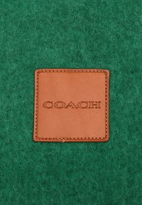 Sfondo in feltro verde con una toppa rettangolare in pelle beige caratterizzata da testo in rilievo "COACH" e cuciture a contrasto.